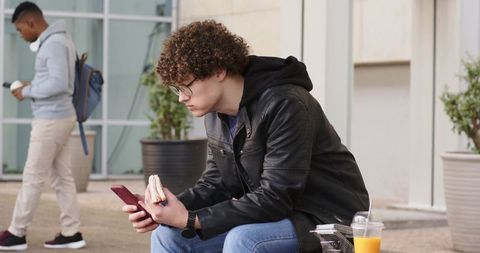Young man in leather jacket eating sandwich while checking smartphone on urban bench