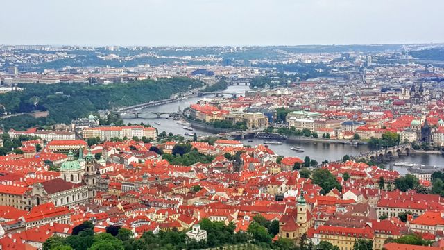 Showcasing an aerial view of Prague, this image overlooks historic landmarks, red-hued rooftops, and scenic Vltava River. The cityscape portrays a blend of historical architecture and modern urbanity, making it perfect for travel guides, tourism promotions, city skyline spotlights, and destination editorials.