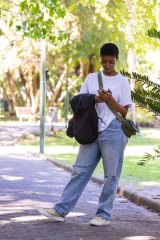 African american female standing in sunlit park checking smartphone, holding jacket