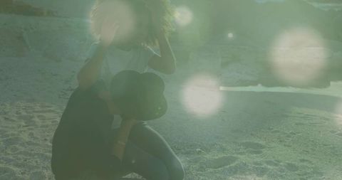 Couple kneeling on sandy shoreline adjusting hair and holding black hat with lens flare