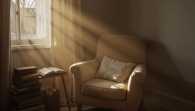 Beige armchair basking in sunbeams beside open book and stacked books