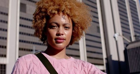 Young Woman Standing in Urban Sunlight Wearing Pink Cable Knit Sweater and Crossbody Bag