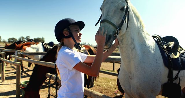 A young Caucasian girl wearing a riding helmet gently touches a white horse's face, expressing affection and trust. Her connection with the animal suggests a passion for equestrian activities and a bond between rider and horse.