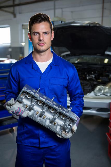 Mechanic in blue overalls holding car engine part in repair garage. Ideal for use in automotive industry promotions, repair service advertisements, and technical training materials.