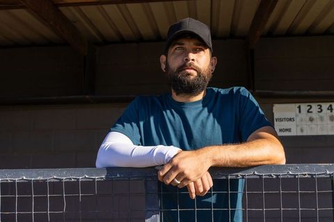 Bearded athlete leaning on dugout fence watching scoreboard in sunlit baseball dugout