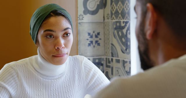 Person of color wearing white sweater and dark green hijab, having serious discussion with another person indoors, in modern and culturally enriched environment. Perfect for use in content related to communication, diversity, cultural interaction, and interpersonal relationships.