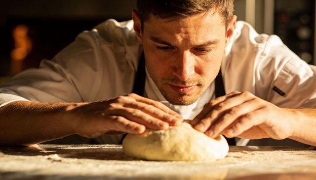 Male baker shaping dough on flour-dusted bench kneading artisan bread with warm oven glow