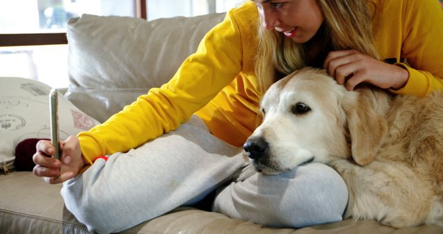 A young woman in casual clothing, wearing a yellow sweater, sits on a couch taking a selfie with her golden retriever. The scene shows a cozy indoor setting, highlighting the joy and bond between pets and their owners. Ideal for use in pet care advertisements, lifestyle blogs, or social media posts focusing on pet ownership and enjoyment at home.