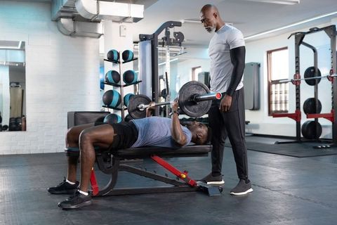 African american trainer spotting man performing barbell bench press on flat bench in gym