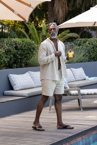 Senior african american man strolling pool deck in linen set holding tropical cocktail