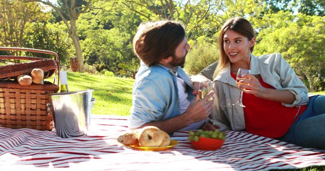 Young couple enjoying a picnic in a park, relaxing on a blanket with wine and snacks. They are surrounded by greenery on a sunny day, creating a perfect outdoor leisure scene. Ideal for use in advertisements for wine brands, outdoor activity promotions, and lifestyle blogs focused on summer activities and relationships.