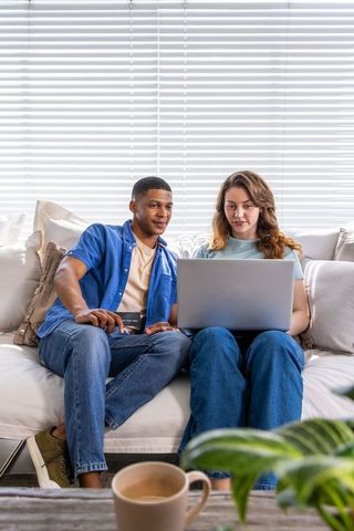 Multiracial couple sitting on sofa sharing laptop and browsing in bright living room
