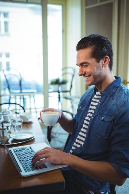 Young man enjoying coffee while working on laptop in a cozy café. Ideal for illustrating remote work, modern lifestyle, technology usage, and casual business environments. Perfect for blogs, articles, and advertisements related to coffee shops, freelancing, and digital nomad lifestyles.