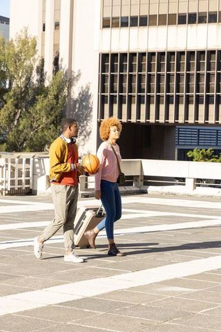 African american couple walking with suitcase and basketball on urban rooftop plaza