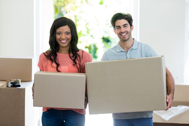 This image features a happy young couple carrying cardboard boxes inside their new home. Ideal for use in real estate advertisements, moving services promotions, and articles about relocation or starting a new home. Highlights themes of teamwork, fresh beginnings, and domestic life.