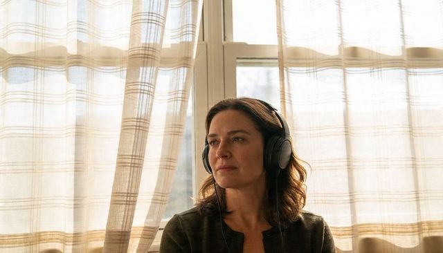 Woman listening with headphones by sunlit window with plaid curtains, contemplative portrait