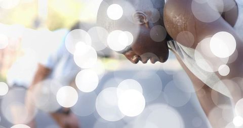 Leaning male athlete catching breath on sunlit track with bokeh highlights and sweat