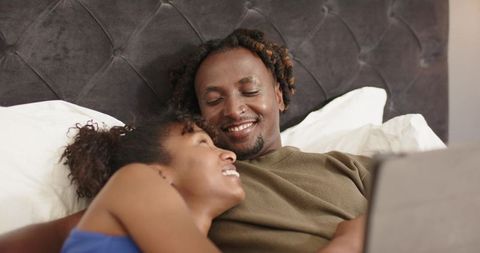 African american couple relaxing in bed sharing laptop, smiling and enjoying cozy moment