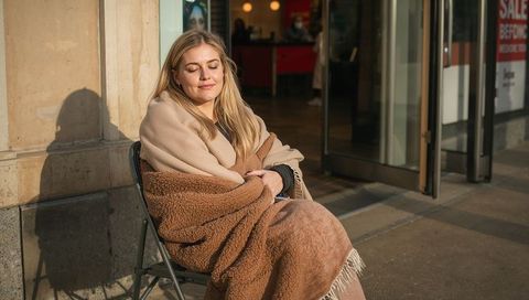 Resting woman wrapped in cozy blanket soaking up sunlight while sitting outside storefront