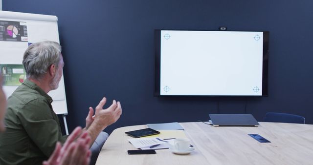 Senior businessman applauding during a presentation meeting in a modern office with a large screen display. Can be used for business and corporate themes, illustrating professional meetings, workplace settings, leadership, and teamwork in a contemporary office environment.