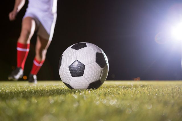 This image captures the lower body of a soccer player in action, focusing on the ball and legs on a grassy field at night. The scene is illuminated by bright lights, highlighting the intensity of the game. Ideal for use in sports-related content, advertisements, and articles about soccer, athleticism, or night games.