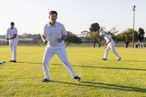 Cricket players practicing fielding drills in white uniforms on sunlit outfield