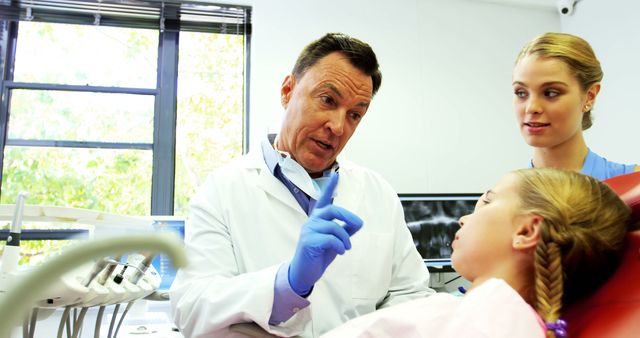 Dentist wearing white coat and blue gloves explaining a dental procedure to a young girl, with a parent present. Useful for topics on healthcare, pediatric dentistry, patient education, dental offices, medical professional services.