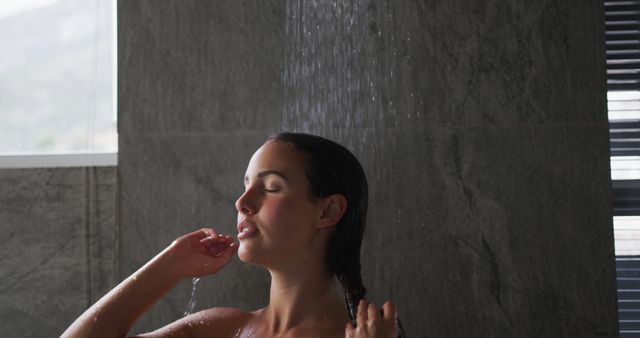 This image shows a woman enjoying a refreshing shower with water droplets falling on her. Ideal for promoting personal hygiene products, relaxation and wellbeing campaigns, or illustrating daily routines. The serene expression and modern bathroom setting evoke a sense of calm and cleanliness.