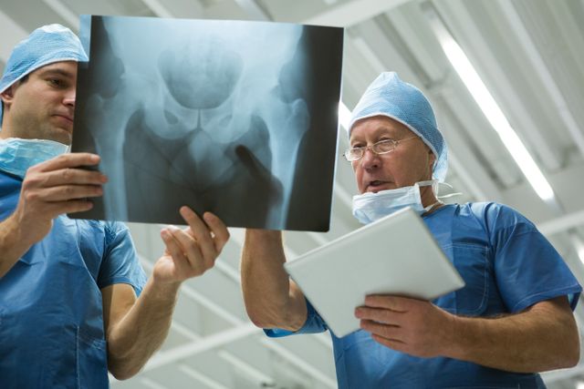 Male surgeons in blue scrubs and surgical caps are discussing an x-ray while holding a digital tablet in a hospital. This image can be used for medical and healthcare-related content, illustrating teamwork, diagnosis, and the use of technology in patient care.