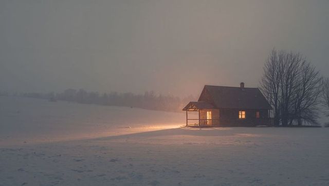 Glowing cabin spilling warm light onto snowy meadow at twilight with bare trees and solitude