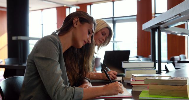 Young women engaged in studying at table stacked with books in modern library with large windows. Ideal for educational and academic content, illustrating student life and focused study environments.