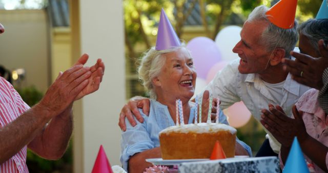 A group of senior Caucasian individuals celebrates a birthday party outdoors, with copy space. Smiles and joy are evident as they gather around a cake with lit candles, sharing a moment of happiness.