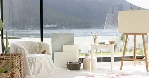 Sunlit coastal art studio with ocean view, white draped armchair, easel and canvases