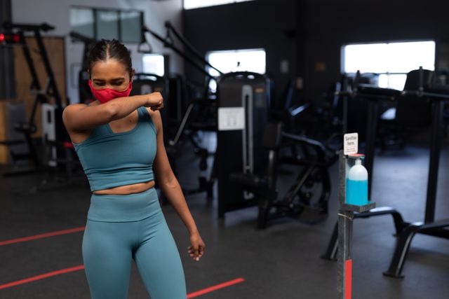Woman sneezing into her arm while wearing a red facemask in a gym. Hand sanitizer bottle nearby. Useful for illustrating health and safety measures in fitness environments, promoting hygiene practices, and emphasizing the importance of wearing masks during workouts.