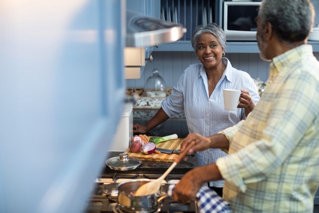 Smiling woman coffee cup talking with man preparing food while standing in kitchen