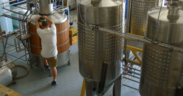 This image shows a brewery worker inspecting large fermentation tanks in a brewery facility. The worker is wearing casual clothes and appears to be thoroughly examining the equipment. This can be used for articles about the brewing industry, brewing processes, industrial photography, or any content related to beer production.