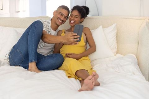 Diverse couple laughing and taking selfie on bed, expecting mother touching belly