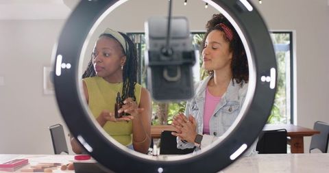 Two diverse women filming beauty tutorial with serum dropper and ring light on marble countertop