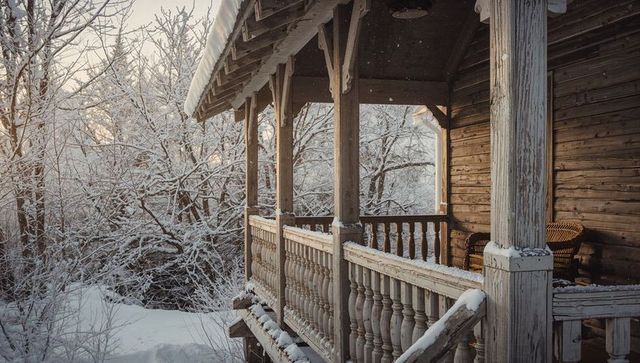 Snow-covered rustic cabin porch with turned spindles and wicker chair at sunrise