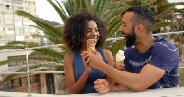 This image captures a happy couple enjoying ice cream cones together outdoors on a pleasant summer day, surrounded by palm trees. Suitable for use in promotions for summer activities, food and beverage advertising, or lifestyle blogs showcasing enjoyable moments and relationships.