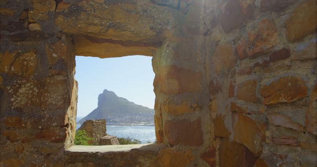 Stone frame from an old, rustic structure opens to a breathtaking view of a coastal mountain with sunlight streaming through. Ideal for travel blogs, historical sites promotion, nature-themed content, and scenic photo collections aimed at invoking a sense of history and natural beauty.