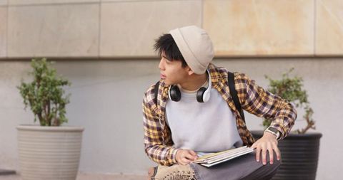Asian male student sitting on ledge looking left with backpack, notebooks and headphones