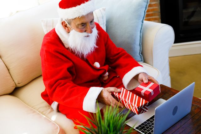Top view of an old caucasian man wearing a santa clause costume holding out a christmas gift on each of his hands while video chatting on a laptop. he is sitting on the couch in the living room.