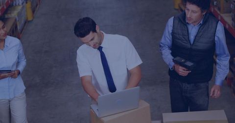 Warehouse supervisor typing on laptop atop boxes while team coordinating inventory and scanning