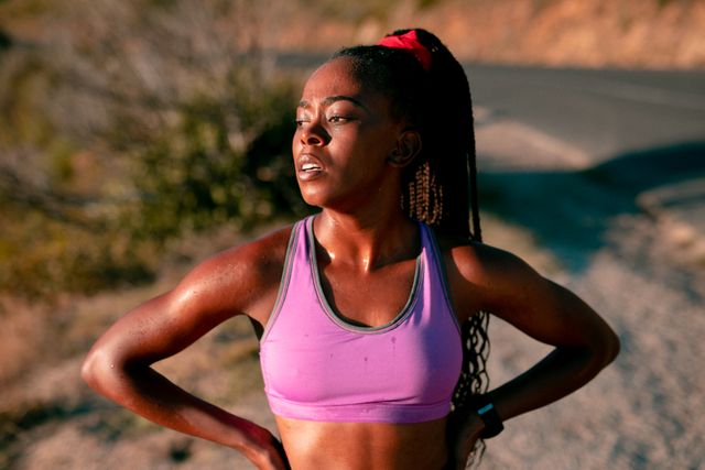 African American woman standing outdoors at sunset, looking determined after a workout. She is wearing athletic wear, including a purple sports bra, and appears sweaty from physical activity. This image is ideal for promoting fitness, healthy lifestyles, outdoor activities, and sportswear. It can be used in articles, advertisements, and social media posts focused on health and wellness.