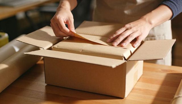Hands Packing Kraft-Wrapped Parcel Into Cardboard Box on Wooden Table