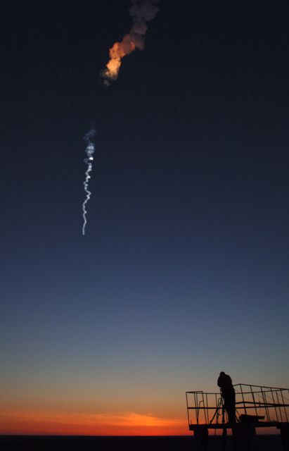 Rocket launching in dramatic dawn sky with silhouette of photographer at Baikonur Cosmodrome in Kazakhstan. Captures moment of Soyuz launch on April 15, 2005, bound for the International Space Station. Useful for articles and marketing materials related to astronomy, space exploration, science education, and space missions.