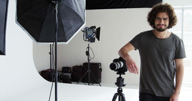 A young Caucasian male photographer stands confidently in his studio, surrounded by professional lighting equipment and a camera on a tripod, with copy space. His casual attire and cheerful demeanor suggest a relaxed yet professional work environment.