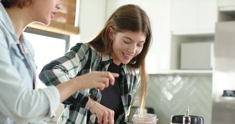 Female friends making smoothie in sunlit kitchen scooping protein powder near blender