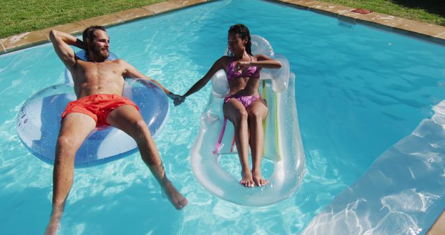 This image depicts a couple enjoying a relaxing time on floats in a swimming pool on a sunny day. The man is lying on a blue float while the woman is on a white float, holding his hand. Ideal for promotions related to summer activities, vacation packages, leisure products, or swimming pool safety materials.